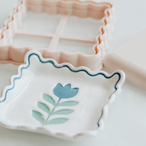 May include: A square ceramic dish with a wavy edge, featuring a blue tulip illustration and a dark blue wavy border. Beside it sits a light pink square plastic cookie cutter with a matching wavy edge and a smooth light pink square block.