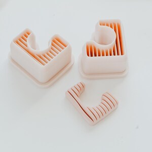 May include: Three light pink, plastic cookie cutters in the shape of the letter "L". Two are larger, with a rectangular base and a curved interior. The third is smaller, with a similar design. The cutters have a ribbed interior.