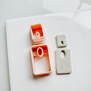 May include: A collection of polymer clay cutters and finished clay pieces. Two pink rectangular cutters with oval cutouts are next to two speckled gray clay rectangles, one with a hole and one with an oval cutout. The items are on a white surface.