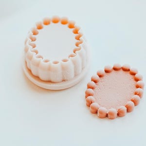 Könnte beinhalten: Eine hellrosa, geriffelte Form und ein passendes ovales Stück mit einem perlenbesetzten Rand. Die Artikel bestehen aus einem festen Material und liegen auf einer weißen Oberfläche. Die Form hat einen kreisförmigen Innenraum.