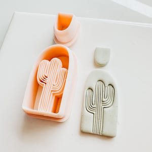 May include: A light pink cactus-shaped polymer clay cutter and a matching clay piece. The cutter has a cactus design with vertical lines. A light green clay piece with a cactus design is also present. The items are on a white surface.