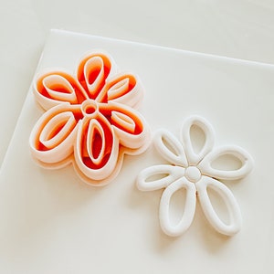 May include: Two flower-shaped cookie cutters, one is orange and the other is white. The orange cutter is slightly larger than the white cutter.