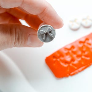 May include: A gray, cylindrical stamp with a bow design is held above a piece of orange clay. Two small, white and yellow objects are in the background. The stamp is likely used for crafting or decorating.