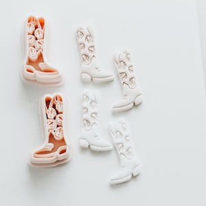 May include: A selection of cowboy boot-shaped cookie cutters and biscuits. The cutters are light pink, and the biscuits are white. The boots have decorative cut-out designs. The boots are arranged on a white surface.