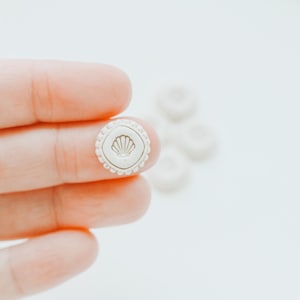 May include: A small, round, off-white button with a seashell design in the centre. The button has a scalloped edge and is held between fingers. Other buttons are blurred in the background. Suitable for sewing and craft projects.