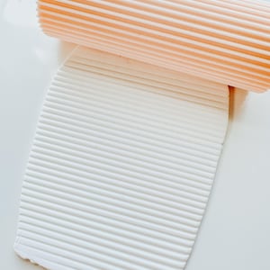 May include: A light orange, ribbed rolling pin rests on a white, textured clay sheet. The clay has been rolled to create a series of parallel lines. The image is shot from a top-down perspective, highlighting the texture and pattern.