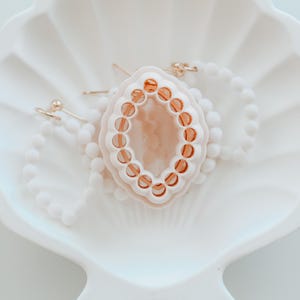 May include: A white, seashell-shaped dish holds a light pink and orange pendant, gold earrings, and a white beaded necklace. The pendant has a decorative, oval design with orange accents. The earrings are small gold studs. The necklace is made of small white beads.