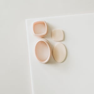 May include: Four pink and beige resin molds for making jewelry. The molds are in the shape of a square, a circle, and two ovals.