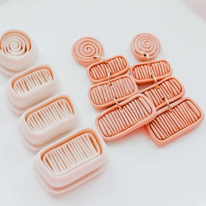 May include: Peach and cream-coloured polymer clay earrings and components. The earrings have a layered rectangular design with linear detailing and a spiral top. The components include a spiral and rectangular shapes. Measurements are in centimetres.