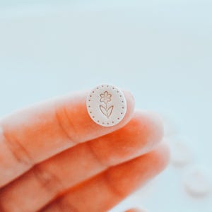 Puede incluir: Un pequeño botón ovalado blanquecino con un diseño floral. El botón presenta una flor con un tallo y hojas en el centro, rodeada por un borde punteado. El botón está sostenido en la mano de una persona.