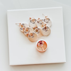 May include: A set of six pairs of gold-tone earrings with white ceramic round dangles featuring a floral design. The earrings are displayed on a white surface.