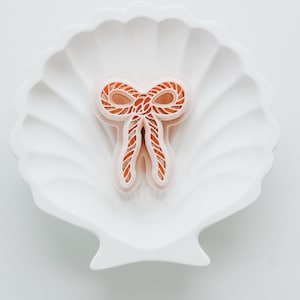 May include: A white, shell-shaped dish holds an orange bow-shaped object. The bow has a rope-like texture and a white outline. The dish has a fluted design and sits on a white surface.