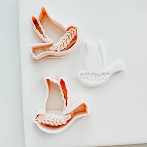 May include: Two pink plastic dove cookie cutters with a leaf design. The cutters are on a white surface.