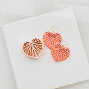 May include: A set of coral-coloured heart-shaped earrings and a matching heart-shaped mould. The earrings have gold-coloured hooks. The heart design features a fan-like pattern. The items are displayed on a white surface.