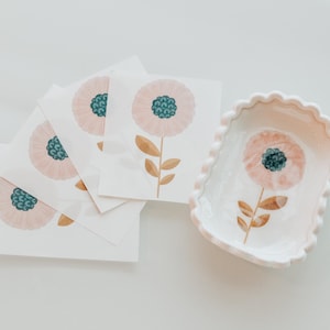 May include: A white ceramic scalloped dish featuring a painted pink flower with a dark teal center and brown stem, accompanied by a stack of four square white cards, each displaying an identical illustration of the same pink flower design.