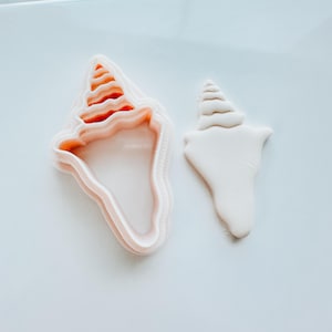 Op de afbeelding: Een schelpvormige koekjesuitsteker en een schelpvormige fondant. De uitsteker is lichtroze met een gedetailleerde rand en binnenkant. De fondant is wit en heeft een vergelijkbare vorm. De items liggen op een wit oppervlak.