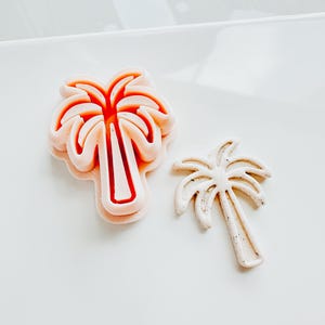 Puede incluir: Dos cortadores de galletas con forma de palmera. Uno es de color rosa claro con el interior rojo, y el otro es de color beige claro. Estos cortadores están diseñados para hornear y crear golosinas con temática tropical.