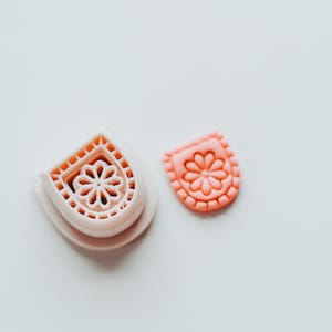 May include: Two pink cookie cutters. One is a floral design within a rounded rectangle, and the other is the matching stamp. The cutters are made of plastic and are on a white surface. The floral design is a flower with six petals.