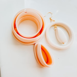May include: A collection of light peach-colored polymer clay earring cutters and a pair of finished earrings with gold-colored hooks. The cutters are in various circular and teardrop shapes. The earrings feature a teardrop-shaped charm.