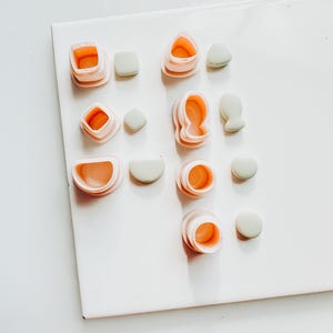 May include: A collection of polymer clay cutters in various shapes, including squares, circles, and teardrops. The cutters are light pink with orange interiors, paired with small, pale green clay pieces. The items are arranged on a white surface.