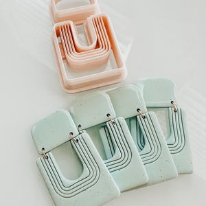 May include: Four pairs of light blue clay earrings with a geometric design. Each earring has a rectangular shape with a curved opening in the center. The earrings are made with a speckled clay and have a subtle texture.