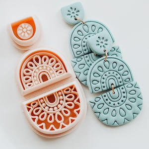 Op de afbeelding: Een paar lichtblauwe, gelaagde oorbellen met een gedetailleerd, halfrond ontwerp en een set bijpassende kleisnijders in perzik. De oorbellen en snijders hebben een herhalend patroon van stippen en bogen.