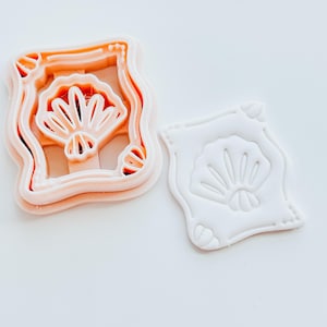 May include: Two cookie cutters. One is peach-colored and has a fan design within a rectangular frame. The other is white and has the same fan design. Both are on a white background. The cutters are likely for baking.