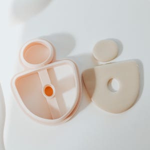 May include: A collection of light pink polymer clay cutters for crafting. The set includes a circular cutter, a half-circle cutter with a hole, and a small circle. The cutters are arranged on a white surface.