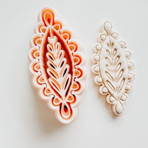 May include: Two leaf-shaped cookie cutters. One is a light peach colour with a detailed leaf design and scalloped edges. The other is a smaller, off-white version with a similar design. Both are on a white surface.