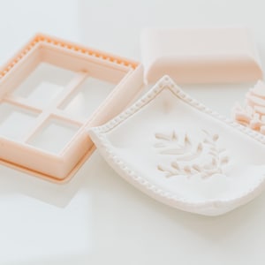 May include: A peach-coloured rectangular window frame cutter, a white curved tray with a raised leaf pattern, a small peach-coloured leaf-shaped stamp, and a smooth peach-coloured rectangular block are arranged on a white surface.