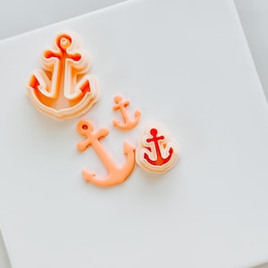 May include: Four orange plastic anchor cookie cutters of different sizes on a white surface.