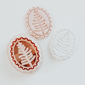 May include: Three oval cookie cutters with a botanical design. One is white, one is light pink, and the third is a darker pink with a brown interior. Each cutter has a scalloped edge and a fern-like leaf design.