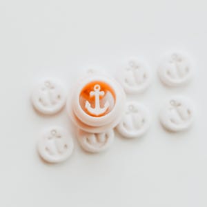 May include: A collection of white wax seals with an anchor design. One seal has an orange center with a white anchor. The seals are arranged on a white surface, creating a nautical theme.