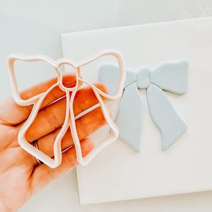 May include: A white plastic cookie cutter in the shape of a bow, held in a hand. A light blue fondant bow is on a white surface behind the cutter.
