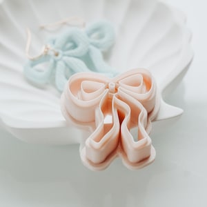 May include: A light pink bow-shaped cookie cutter is the focus, with a light blue bow in the background. Both are on a white, shell-shaped dish. The bow-shaped cutter is for baking. The bow is made of fabric.