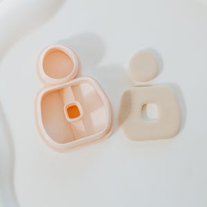 May include: Two light peach-colored polymer clay earring cutters and a finished earring. One cutter is a rounded rectangle with a small square cutout, the other is a circle and a rounded rectangle with a square cutout.