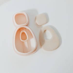 May include: A collection of polymer clay cutters and clay pieces in a soft peach color. The cutters are in various organic shapes, including a square, a teardrop, and an oval with a hole. The clay pieces are in matching shapes.