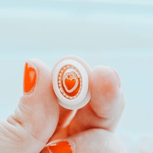 May include: A small, white and orange button with a heart design. The button has a layered design with a heart in the centre and a dotted border. The background is a light blue.