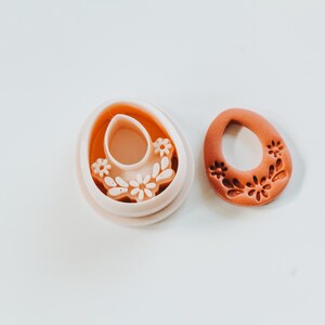 May include: A light orange polymer clay earring and a matching cutter. The earring has a floral design. The cutter is egg-shaped with a floral design and a centre cutout.