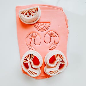 May include: A set of four white plastic cookie cutters in the shape of a lemon wedge and two prawns. The cutters are on top of a pink clay surface.
