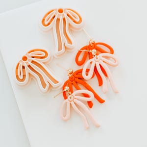 Op de afbeelding: Een paar statement oorbellen met een gelaagd ontwerp, een grote, abstracte bloemenvorm in wit en een kleinere, abstracte strik in oranje en roze.