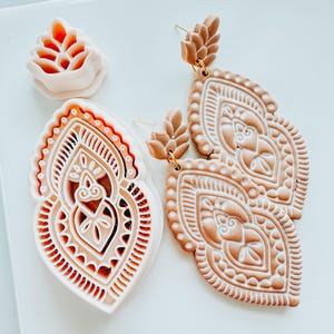 May include: Tan and white clay earrings with a detailed, ornate design. The earrings are in a teardrop shape with a floral accent at the top. A matching clay cutter and a floral clay stamp are also visible.