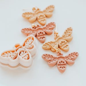 May include: A selection of butterfly-shaped earrings and a cutter. The earrings are in peach and tan hues, with detailed embossed patterns. The cutter is light pink. The earrings have gold-coloured hooks.