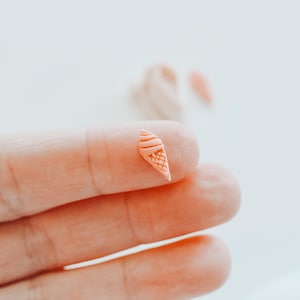 Könnte beinhalten: Eine pfirsichfarbene Miniatur-Eistütenfigur mit einem strukturierten Waffelmuster auf der Tüte und einer spiralförmigen Oberseite. Das kleine Objekt ist detailreich gefertigt und zeigt eine klassische Dessertform in einem hellen Orangeton.