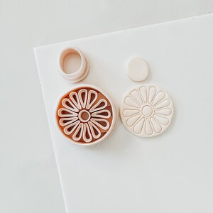 May include: A set of three pink plastic cookie cutters in the shape of a flower. The cutters are on a white surface.