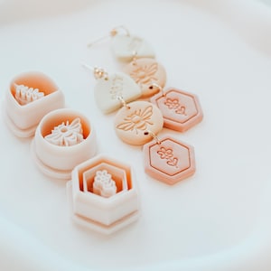 May include: Handmade earrings and clay molds with bee and flower designs. The earrings have gold-coloured hooks and are made of beige, peach, and pink clay. The molds are shaped like a hexagon, circle, and V-shape. The earrings are a unique accessory.
