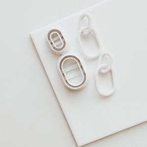 May include: Three white plastic cookie cutters shaped like oval links of a chain. The cutters are on a white surface.