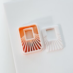 May include: Two-piece set of light pink and orange cookie cutters. One cutter is a rectangular frame with a sunburst design, and the other is a matching embossed shape. The cutters are made of plastic and are designed for baking.