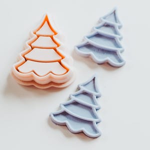May include: Three Christmas tree-shaped cookie cutters. One is a light peach color, and the other two are light blue. The cutters are made of plastic and are on a white surface. The tree shapes have layered, scalloped edges.