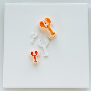May include: A set of four cookie cutters in the shape of a lobster claw. The cutters are made of white plastic and have a red and orange color scheme.